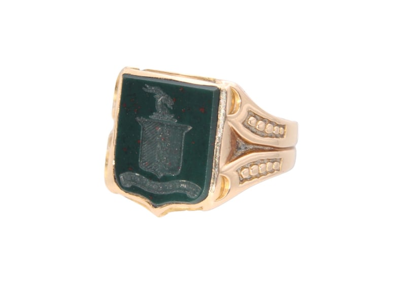 May include: A gold ring featuring a rectangular dark green stone inlay. The stone is engraved with a crest design depicting a shield, a small animal figure at the top, and a banner at the bottom. The gold band has textured side detailing.
