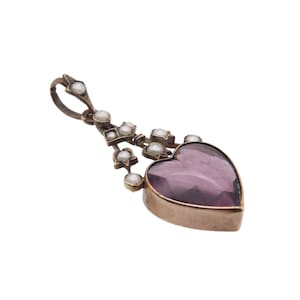 Puede incluir: Un colgante vintage con una piedra preciosa morada en forma de coraz&oacute;n. El colgante est&aacute; adornado con peque&ntilde;as perlas redondas y formas geom&eacute;tricas, suspendidas de un lazo. El metal tiene un tono dorado.