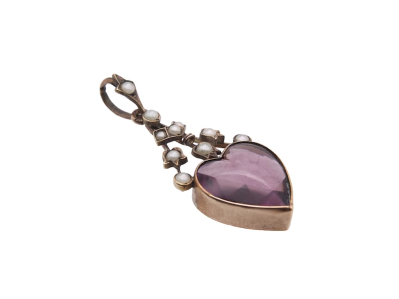 Puede incluir: Un colgante vintage con una piedra preciosa morada en forma de coraz&oacute;n. El colgante est&aacute; adornado con peque&ntilde;as perlas redondas y formas geom&eacute;tricas, suspendidas de un lazo. El metal tiene un tono dorado.