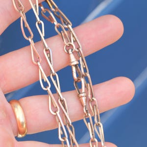 May include: A rose gold-toned metal chain necklace featuring elongated, teardrop-shaped links. The chain includes a small, integrated swivel clasp mechanism for securing the ends together, showcasing a metallic finish with a warm, pinkish hue.