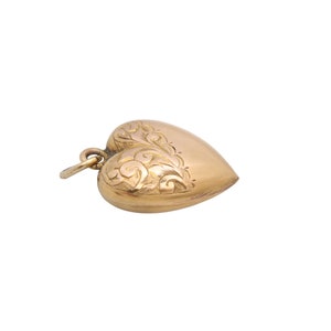May include: A gold-colored heart-shaped pendant with intricate floral engravings on one side. The pendant has a small loop at the top for attaching to a chain. The heart is slightly rounded and has a polished finish.