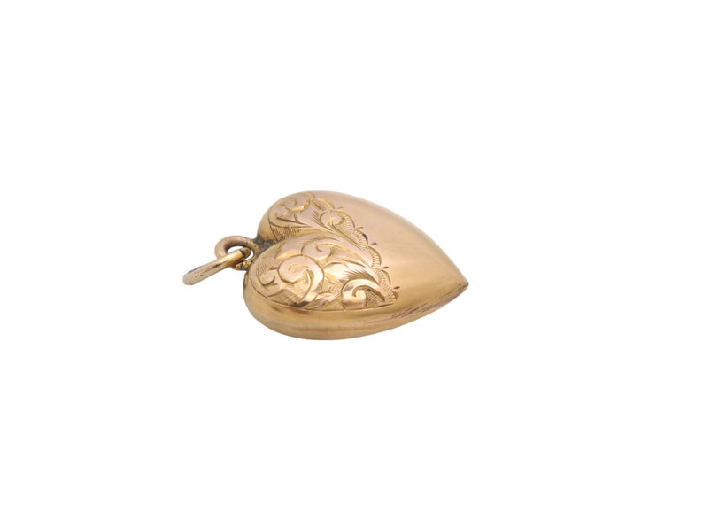 May include: A gold-colored heart-shaped pendant with intricate floral engravings on one side. The pendant has a small loop at the top for attaching to a chain. The heart is slightly rounded and has a polished finish.