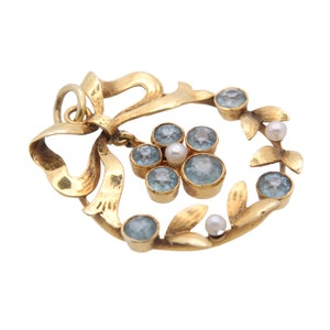 May include: A gold-toned pendant featuring a bow and a wreath design. The wreath is adorned with light blue gemstones and small pearls. The pendant has a loop for hanging.