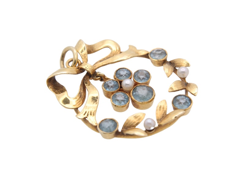 May include: A gold-toned pendant featuring a bow and a wreath design. The wreath is adorned with light blue gemstones and small pearls. The pendant has a loop for hanging.