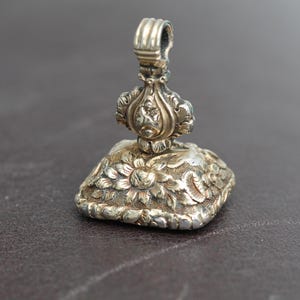 May include: A silver antique wax seal with intricate floral and foliate designs. The seal features a square base with a detailed flower pattern and a decorative handle. The metal has a tarnished appearance, suggesting age and use.