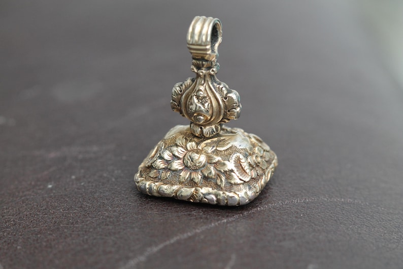 May include: A silver antique wax seal with intricate floral and foliate designs. The seal features a square base with a detailed flower pattern and a decorative handle. The metal has a tarnished appearance, suggesting age and use.