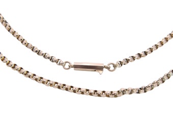 May include: A silver-colored necklace with a box chain design. The necklace features a rectangular clasp in the center, and the chain is composed of small, interconnected square links. The background is plain white.