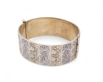 Puede incluir: Pulsera plateada con bisagras y diseño floral grabado. La pulsera presenta paneles alternos de plata y metal dorado, creando un contraste llamativo. El cierre es visible en el lateral. Una pieza de joyería clásica.