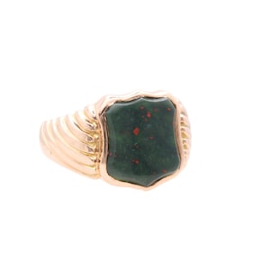 Puede incluir: Un anillo de oro con una piedra verde rectangular en forma de escudo con motas rojas. La banda del anillo tiene un diseño acanalado. El anillo está sobre un fondo blanco.