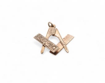 May include: A gold-colored pendant featuring a Masonic symbol. The pendant includes a compass, a square, and a ruler, all with intricate engraved details. The pendant has a small loop for attaching to a chain or cord.