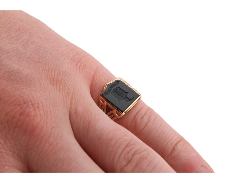 May include: A gold signet ring featuring a rectangular black onyx inlay with a shield-shaped crest engraved on the surface. The metal band displays intricate cut-out detailing along the sides, providing a structured and ornate appearance.