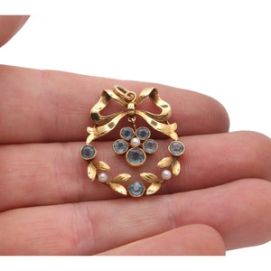 May include: A gold-toned pendant featuring a wreath design with blue gemstones and small pearls. The pendant has a bow at the top and leaf accents. The jewelry is held in a hand against a white background.