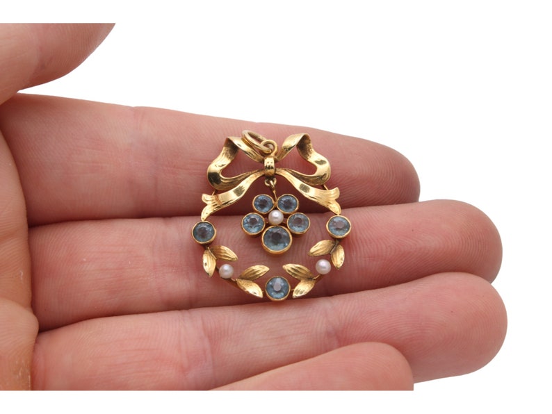 May include: A gold-toned pendant featuring a wreath design with blue gemstones and small pearls. The pendant has a bow at the top and leaf accents. The jewelry is held in a hand against a white background.