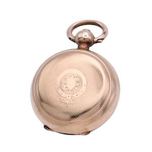 May include: A gold-toned pocket watch with a circular face and a textured, engraved design. The watch features a loop at the top for a chain and an ornate, circular emblem on the front. The watch has a classic, vintage aesthetic.