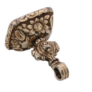 May include: Antique gold-colored seal with floral and leaf detailing. The seal's top features a rectangular design with a prominent sunflower and other flowers. The handle has a decorative design and a loop.
