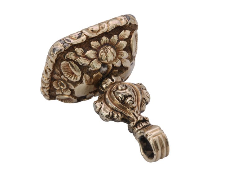 May include: Antique gold-colored seal with floral and leaf detailing. The seal's top features a rectangular design with a prominent sunflower and other flowers. The handle has a decorative design and a loop.