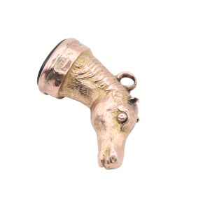 May include: A gold-colored pendant in the shape of a horse's head. The horse head is detailed with a mane, eye, and ear. The pendant has a dark, possibly black, stone at the top and a small loop for a chain.