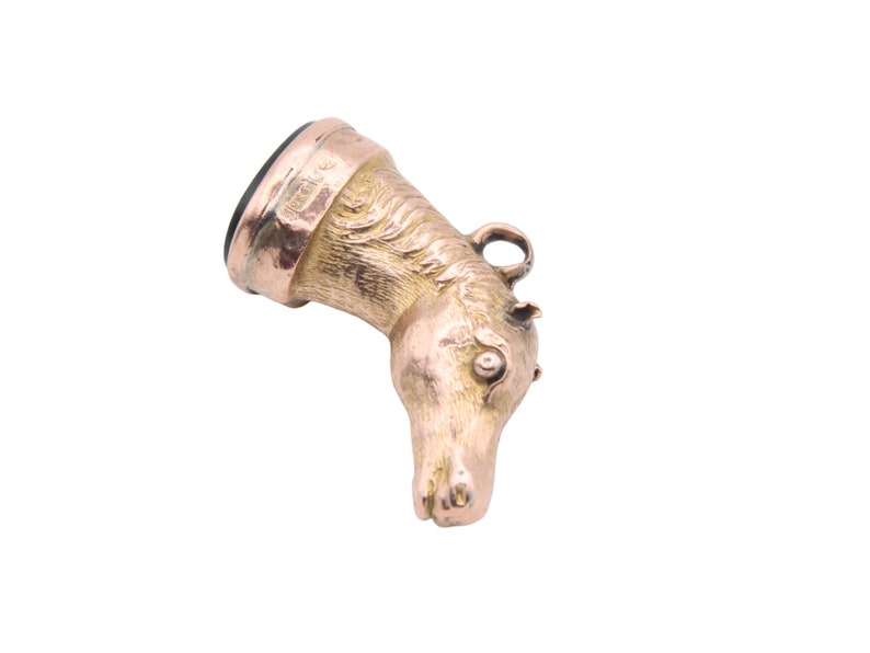 May include: A gold-colored pendant in the shape of a horse's head. The horse head is detailed with a mane, eye, and ear. The pendant has a dark, possibly black, stone at the top and a small loop for a chain.