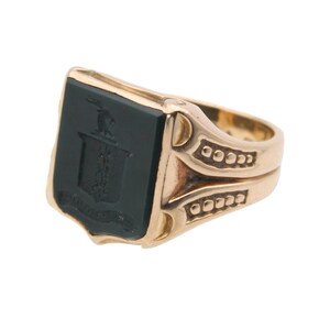 May include: A gold signet ring featuring a rectangular black onyx inlay. The black stone is engraved with a crest design depicting a small animal figure above a shield and a banner. The gold band has decorative side detailing with small raised dots.