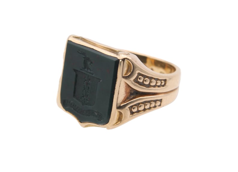 May include: A gold signet ring featuring a rectangular black onyx inlay. The black stone is engraved with a crest design depicting a small animal figure above a shield and a banner. The gold band has decorative side detailing with small raised dots.