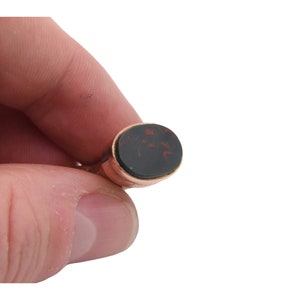 May include: A gold-toned ring with an oval, dark red and black stone. The ring is held between fingers against a white background. The stone has a unique pattern.