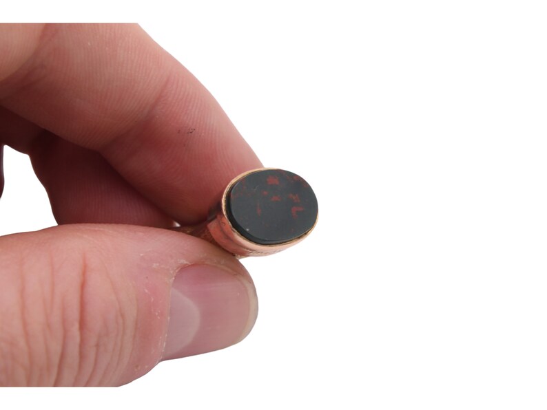 May include: A gold-toned ring with an oval, dark red and black stone. The ring is held between fingers against a white background. The stone has a unique pattern.