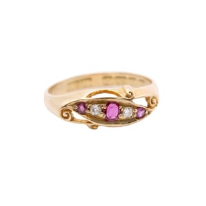 May include: A gold ring featuring a row of gemstones. The ring has a central oval ruby flanked by two smaller rubies and two clear stones. The band is a polished gold with decorative swirls on the face.