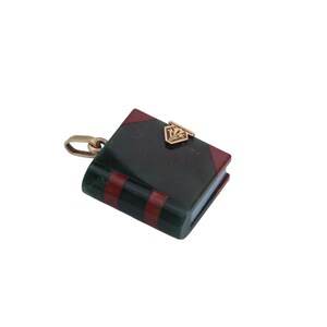 May include: A miniature book charm with a gold-colored clasp and chain. The book has a dark green cover with red stripes and red corners. A gold-colored emblem is on the front cover.