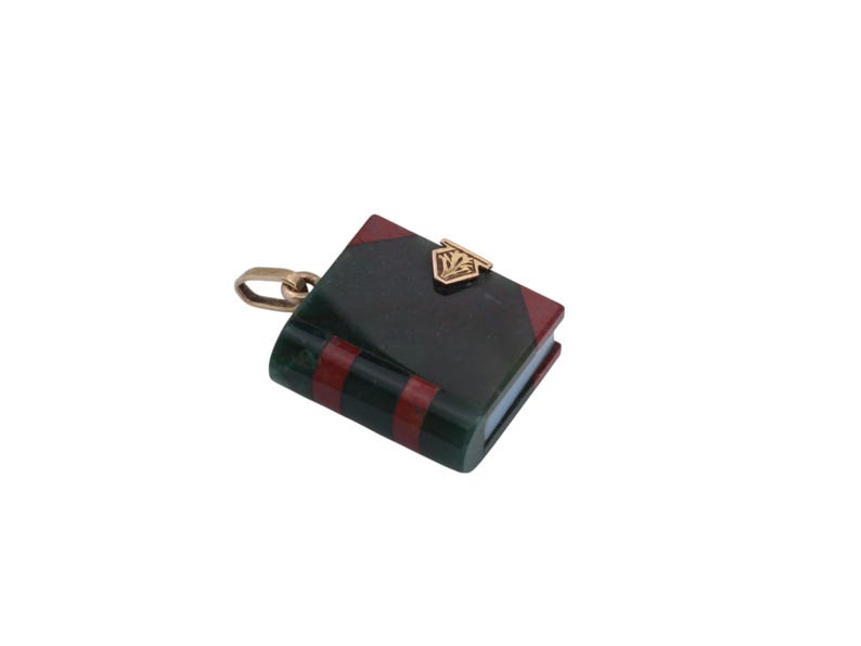 May include: A miniature book charm with a gold-colored clasp and chain. The book has a dark green cover with red stripes and red corners. A gold-colored emblem is on the front cover.