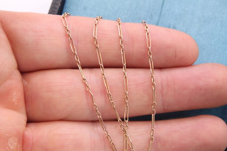 K&ouml;nnte beinhalten: Nahaufnahme einer zarten Goldhalskette mit einem Paperclip-Kettendesign. Die Halskette wird vor einem hellblauen Hintergrund pr&auml;sentiert. Die Kette besteht aus kleinen, rechteckigen Gliedern.
