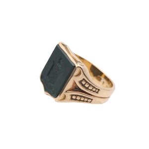 May include: A gold ring featuring a rectangular black stone inlay on the face. The stone is engraved with a crest design. The band has decorative cutouts and small square indentations along the sides, providing a textured appearance.
