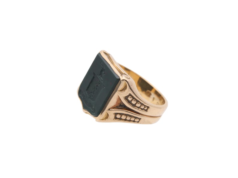 May include: A gold ring featuring a rectangular black stone inlay on the face. The stone is engraved with a crest design. The band has decorative cutouts and small square indentations along the sides, providing a textured appearance.