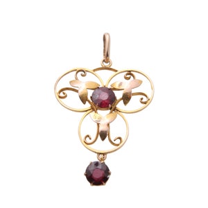 May include: A gold-toned pendant with a floral design and three circular elements. The pendant features three round, deep red gemstones and a smaller, dangling gemstone at the bottom. The design includes leaf-shaped accents.