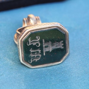 May include: Antique gold-toned signet ring with a dark green, octagonal face. The face features engraved initials and a castle design. The ring is set against a blue background, showcasing its intricate details and vintage aesthetic.