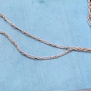 K&ouml;nnte beinhalten: Eine zarte, goldfarbene Halskette mit einem Design aus l&auml;nglichen Gliedern. Die Halskette wird auf einer hellblauen Oberfl&auml;che pr&auml;sentiert, m&ouml;glicherweise in einer Schmuckschatulle. Die Kettenglieder sind gleichm&auml;&szlig;ig angeordnet.