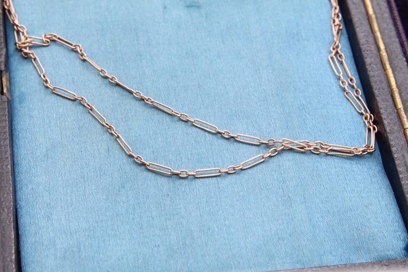 K&ouml;nnte beinhalten: Eine zarte, goldfarbene Halskette mit einem Design aus l&auml;nglichen Gliedern. Die Halskette wird auf einer hellblauen Oberfl&auml;che pr&auml;sentiert, m&ouml;glicherweise in einer Schmuckschatulle. Die Kettenglieder sind gleichm&auml;&szlig;ig angeordnet.
