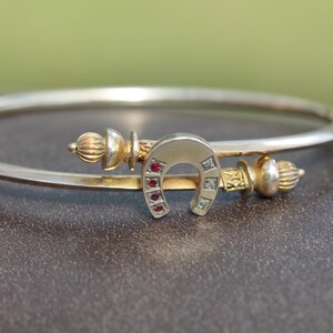 Könnte beinhalten: Ein silberfarbenes Armreif-Armband mit einem goldfarbenen Hufeisen-Anhänger. Das Hufeisen ist mit kleinen roten und klaren Edelsteinen besetzt. Das Armband hat goldfarbene Zierenden.