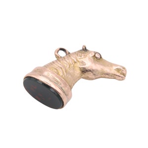 May include: A gold-toned horse head pendant with a dark red, rectangular base. The horse head is detailed with textured fur and a small loop at the top. The base has engraved text.