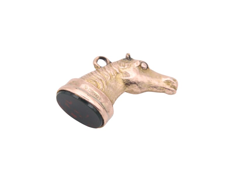 May include: A gold-toned horse head pendant with a dark red, rectangular base. The horse head is detailed with textured fur and a small loop at the top. The base has engraved text.