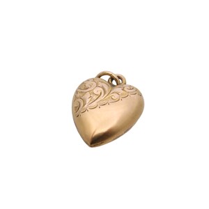 May include: A gold-colored heart-shaped pendant with an ornate, engraved floral design along the top edge. The pendant has a rounded, three-dimensional form and a small loop for a chain.
