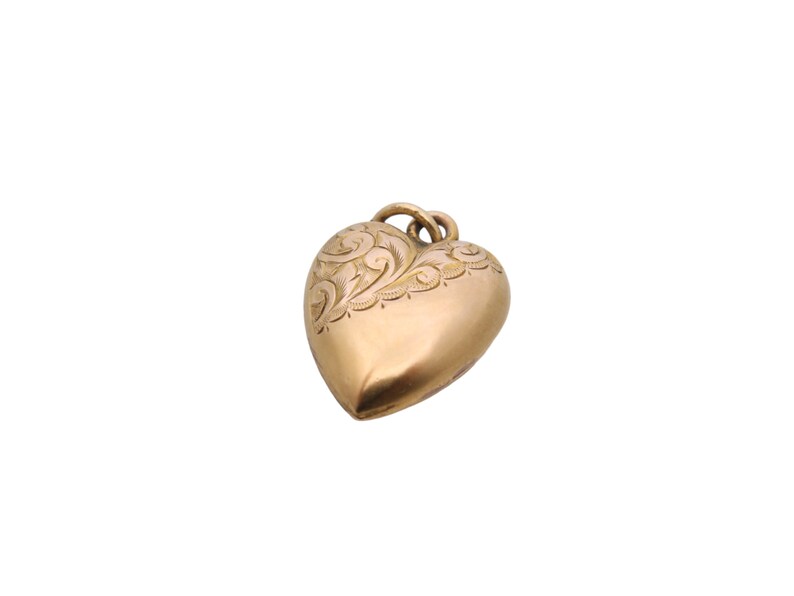 May include: A gold-colored heart-shaped pendant with an ornate, engraved floral design along the top edge. The pendant has a rounded, three-dimensional form and a small loop for a chain.