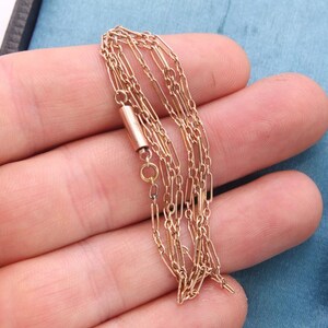 K&ouml;nnte beinhalten: Eine zarte goldfarbene Halskette mit mehreren Str&auml;ngen, gehalten in einer Hand. Die Halskette hat einen kleinen zylindrischen Verschluss und feine Kettenglieder. Der Hintergrund umfasst eine blaue Oberfl&auml;che und eine dunkel gef&auml;rbte Box.
