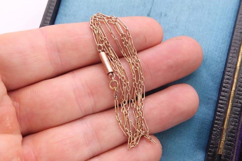 K&ouml;nnte beinhalten: Eine zarte goldfarbene Halskette mit mehreren Str&auml;ngen, gehalten in einer Hand. Die Halskette hat einen kleinen zylindrischen Verschluss und feine Kettenglieder. Der Hintergrund umfasst eine blaue Oberfl&auml;che und eine dunkel gef&auml;rbte Box.