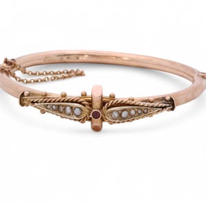 May include: A gold-coloured hinged bangle bracelet. The bracelet has a central decorative section with seed pearls, a small red gemstone, and rope detailing. A delicate chain secures the clasp.