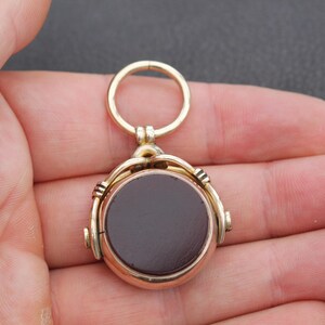 K&ouml;nnte beinhalten: Ein goldfarbener Anh&auml;nger mit einem dunkelbraunen, kreisf&ouml;rmigen Mittelteil. Der Anh&auml;nger hat oben einen Ring zum Aufh&auml;ngen und wird in einer Hand gehalten. Der Anh&auml;nger hat ein Vintage- oder antikes Aussehen.