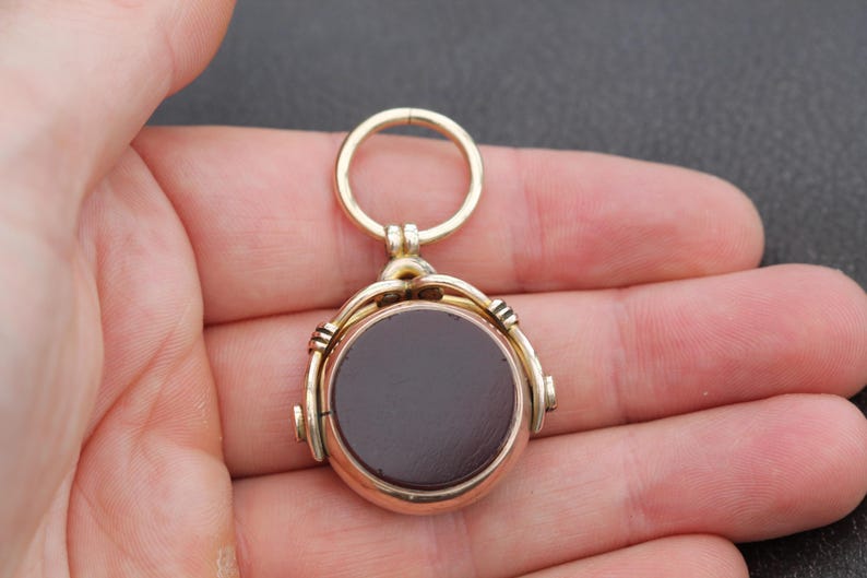 K&ouml;nnte beinhalten: Ein goldfarbener Anh&auml;nger mit einem dunkelbraunen, kreisf&ouml;rmigen Mittelteil. Der Anh&auml;nger hat oben einen Ring zum Aufh&auml;ngen und wird in einer Hand gehalten. Der Anh&auml;nger hat ein Vintage- oder antikes Aussehen.