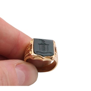 May include: A gold signet ring featuring a rectangular dark green stone inlay. The stone is engraved with a crest depicting a bird above a shield and a banner. The band of the ring has decorative cutouts and a polished metallic finish.