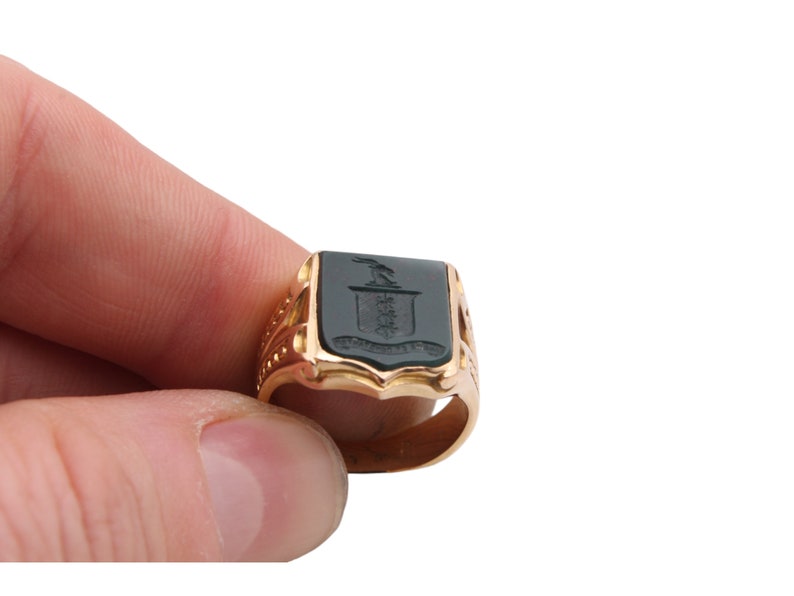 May include: A gold signet ring featuring a rectangular dark green stone inlay. The stone is engraved with a crest depicting a bird above a shield and a banner. The band of the ring has decorative cutouts and a polished metallic finish.
