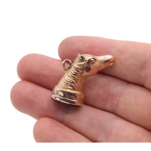May include: A gold-colored pendant in the shape of a horse's head. The horse head is detailed with textured fur and a small loop for a chain. The pendant is held in a person's hand against a white background.
