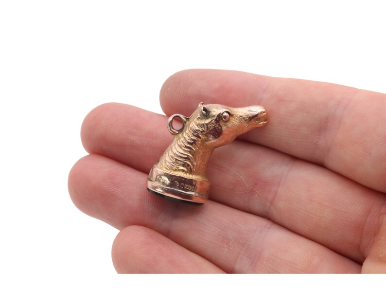 May include: A gold-colored pendant in the shape of a horse's head. The horse head is detailed with textured fur and a small loop for a chain. The pendant is held in a person's hand against a white background.